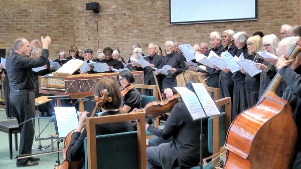 A choir performing Handel's Messiah, with a director leading from the front, surrounded by singers holding sheet music and musicians playing string instruments.