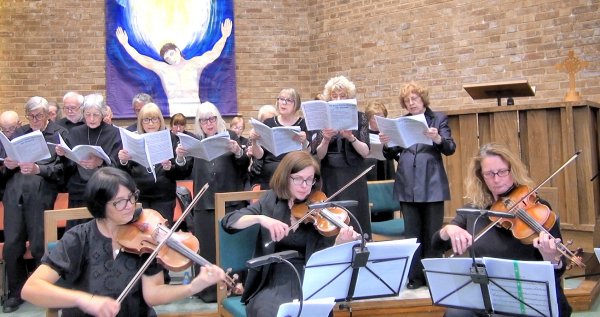 A choir performs indoors, with several singers holding music sheets. In the foreground, musicians play violins. A religious painting is visible in the background.